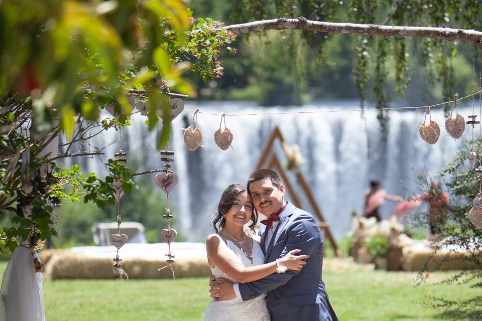 Novios abrazados en un jardín con una cascada de fondo